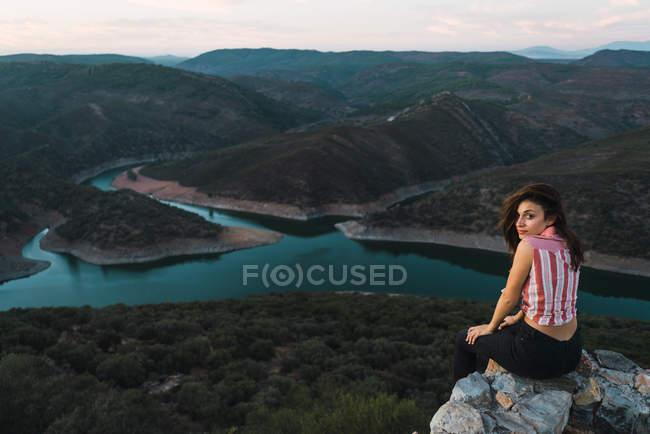 Junge Brünette posiert auf Klippe Kante und blickt über die Schulter in die Kamera über atemberaubenden Hintergrund von Bergen und Flusstal — Stockfoto