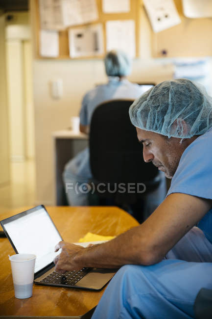 Vista lateral del médico sentado en el ordenador portátil y hablando por teléfono - foto de stock