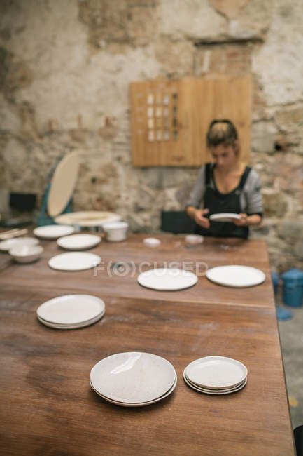 Placas artesanais brilhantes na mesa sobre oleiro feminino trabalhando com argila na oficina — Fotografia de Stock