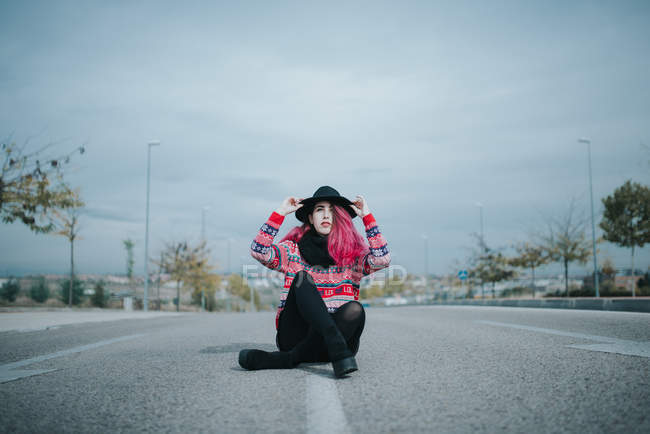 Pretty young girlin jumper sitting on road and adjusting hat. — Stock Photo