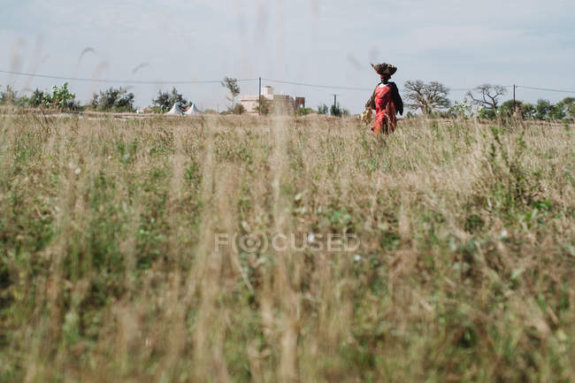Yoff, Senegal- December 6, 2017: Woman walking on sunny field and carrying basket on head. — Stock Photo