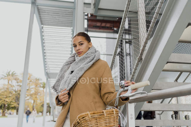 High angle portrait of young woman with city bicycle wrapping in coat and looking at camera — Stock Photo