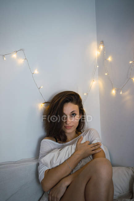Брюнетка сидит на кровати за стеной со сказочными огнями и смотрит в камеру — стоковое фото