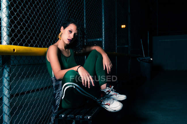 Vue latérale de la fille assise sur le sol par une clôture métallique et regardant la caméra — Photo de stock