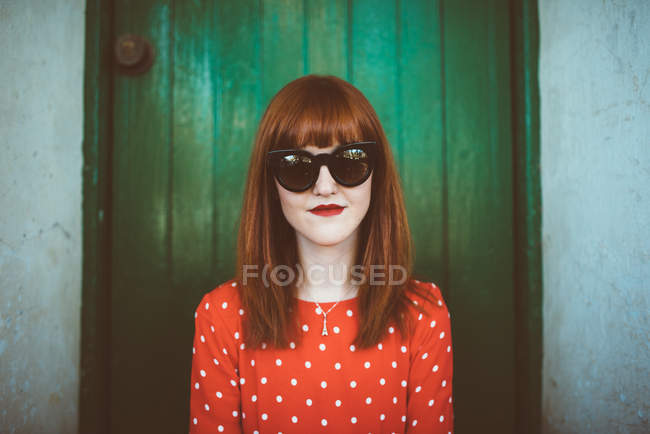 Retrato de mujer pelirroja en ropa roja y gafas de sol posando sobre pared de madera verde - foto de stock