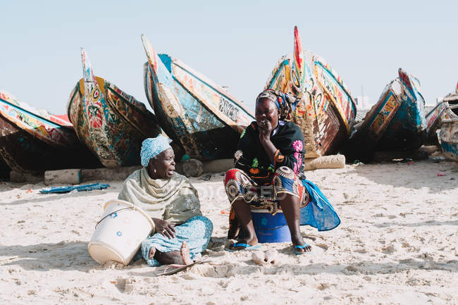 Yoff, Senegal- December 7, 2017: Two women sitting and smiling by old vessels on sunny sandy coast. — Stock Photo