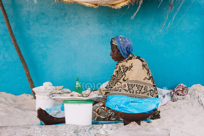 Yoff, Senegal- December 6, 2017: Side view of black old lady sitting barefoot on street pavement against bright wall with some goods. — Stock Photo