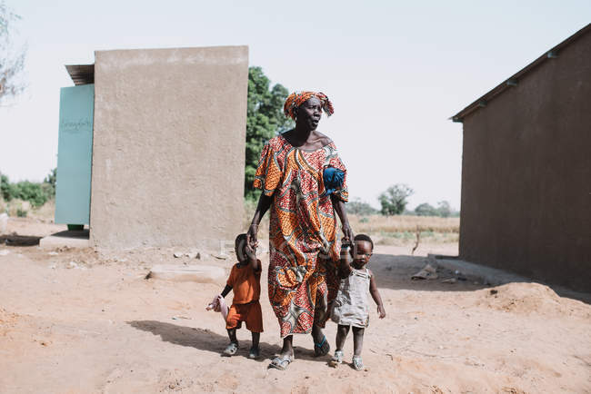 Goree, senegal- 6. Dezember 2017: Afrikanerin hält Hände von Kleinkindern, während sie auf dem Sand der Straße in der armen Landschaft läuft. — Stockfoto