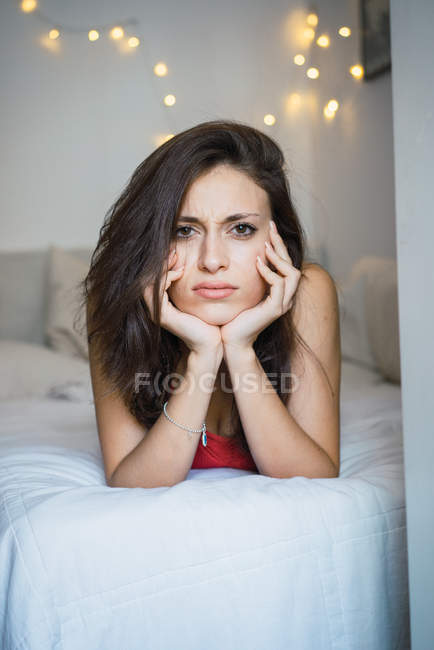 Unzufriedenes brünettes Mädchen, das mit dem Kopf auf den Händen auf ihrem Bett liegt und in die Kamera schaut. — Stockfoto