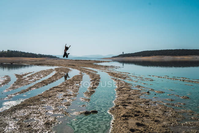 Віддалений вигляд жінки, що стрибає через брудну місцевість з озерною водою — стокове фото