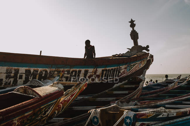 Yoff, Sénégal- 6 décembre 2017 : Un Africain assis sur un bateau naufragé peint sur le rivage avec de vieux voiliers en pleine lumière
. — Photo de stock