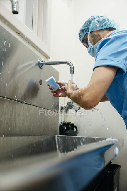 Боковой вид человека в синей медицинской форме с помощью губки при тщательном мытье рук — стоковое фото
