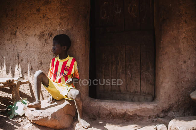 Goree, Senegal- Dezembro 6, 2017: Menino descalço sentado no alpendre de pedra da casa da aldeia pobre  . — Fotografia de Stock