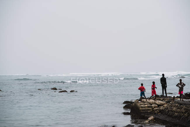 Goree, Senegal - 6 dicembre 2017: Gruppo di persone africane in piedi su una piccola roccia vicino al mare che ondeggia . — Foto stock