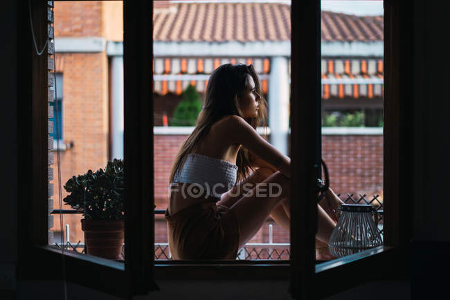 Vue latérale de la fille brune se détendre sur le rebord de la fenêtre à la terrasse de l'appartement — Photo de stock