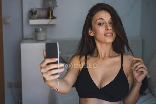 Brünettes Mädchen in schwarzem BH macht Selfie im Schlafzimmer. — Stockfoto