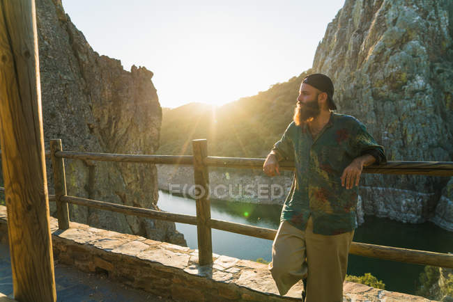 Bearded man leaning on terrace fence over view of mountain river — Stock Photo