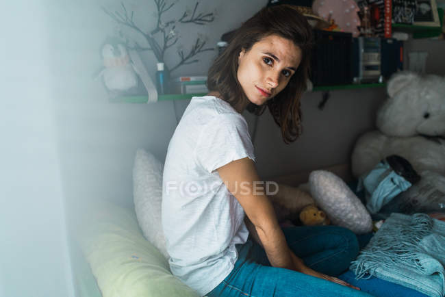 Morena menina sentada na cama entre almofadas e brinquedos e olhando para a câmera . — Fotografia de Stock