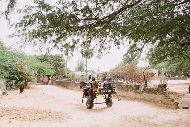 Goree, Senegal- December 6, 2017: Group of black children on cart harnessed with horse riding down road in village. — Stock Photo