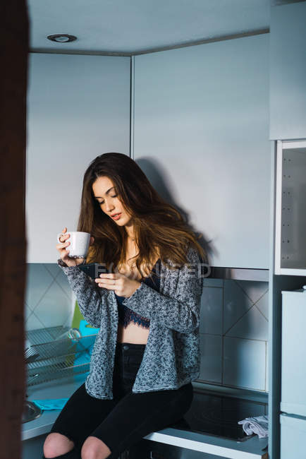 Chica morena sentada en el mostrador de la cocina con taza de café y teléfono inteligente - foto de stock