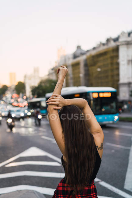 Брюнетка с видом сзади позирует с поднятыми руками рядом с городской дорогой — стоковое фото