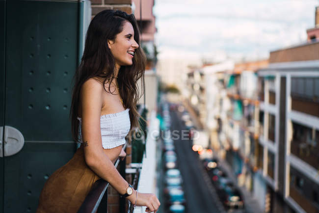 Вид сбоку улыбающейся брюнетки, облокотившейся на балкон через забор на улице — стоковое фото