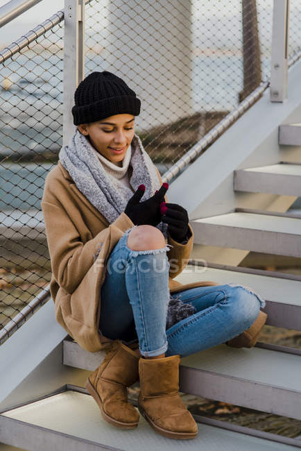 Mujer sonriente sentada en la escalera y el teléfono inteligente de navegación - foto de stock