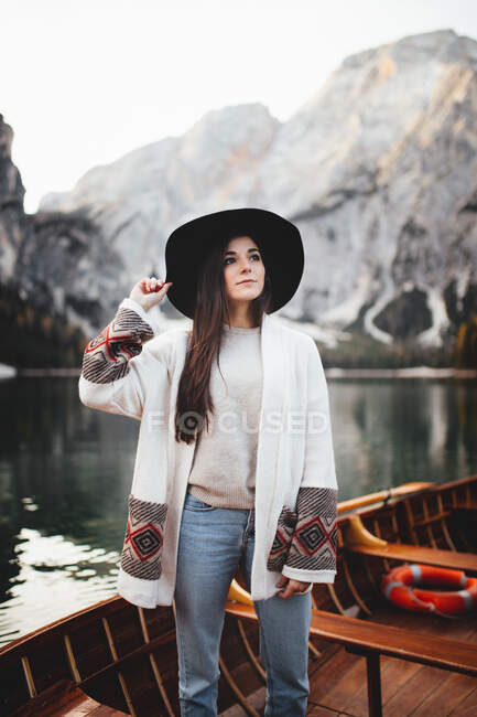 Bella giovane donna in cappello e giacca in piedi in barca sullo sfondo di belle montagne. — Foto stock