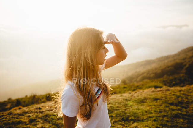 Pretty young woman standing in nature, holding her hand near her forehead and looking into distance. — Stock Photo