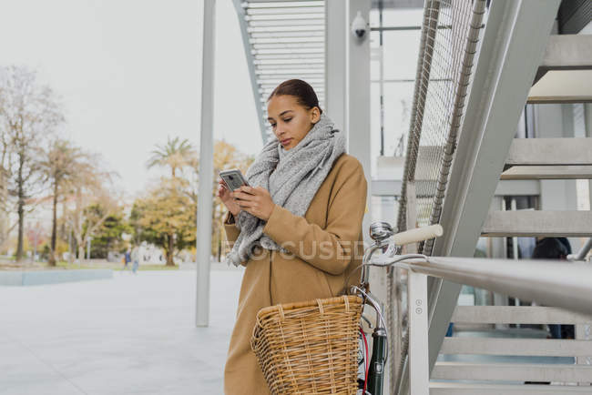 Mulher bonita de casaco e de pé perto de bicicleta estacionada e browsingphone — Fotografia de Stock