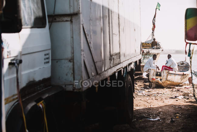 Goree, Sénégal- 6 décembre 2017 : Vue lointaine d'hommes assis sur des bateaux à la plage près de camions garés et parlant — Photo de stock