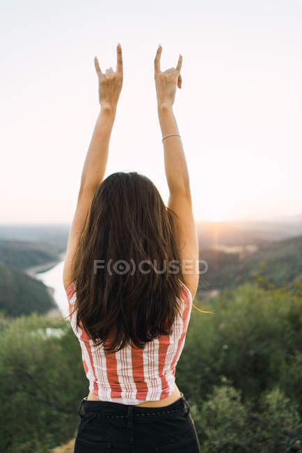 Rear view of girl posing with raised arms and rock gesture on background of mountain valley landscape — Stock Photo