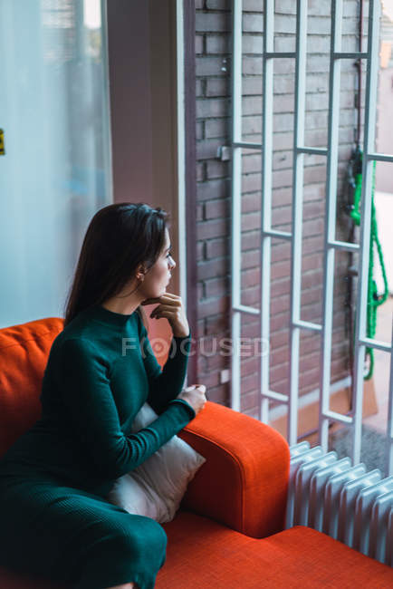 Вид сбоку женщины в платье, позирующей на красном диване и задумчиво смотрящей в окно . — стоковое фото