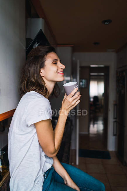 Vista laterale della ragazza bruna seduta sul bancone della cucina e che beve caffè — Foto stock