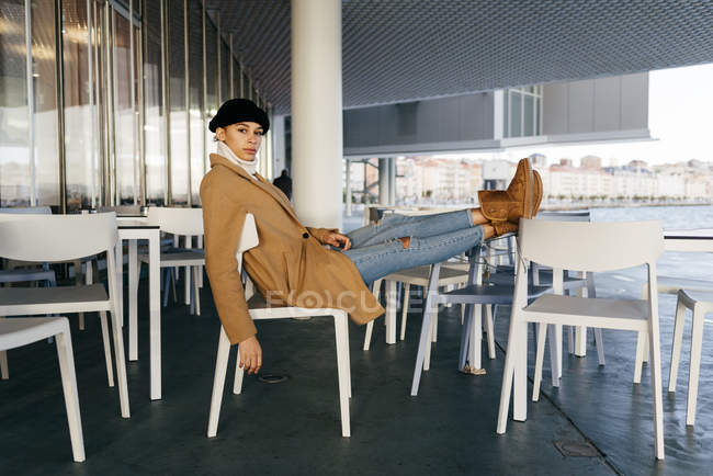 Vista lateral da mulher em roupas quentes com os pés na mesa de café olhando para a câmera — Fotografia de Stock