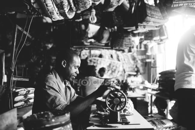 Goree, Senegal 6 de diciembre de 2017: Vista lateral del hombre sentado usando una vieja máquina de coser en la mesa en el taller y  . - foto de stock