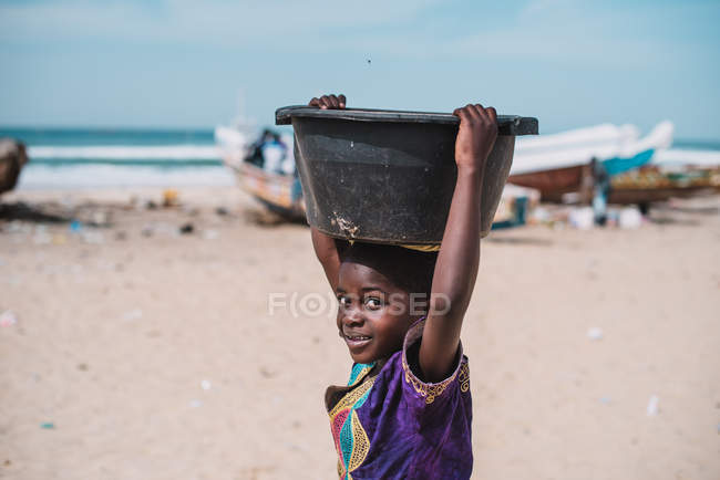 Goree, senegal- 6. Dezember 2017: Porträt eines Mädchens, das Plastikbecken auf dem Kopf trägt und am Strand in die Kamera blickt — Stockfoto
