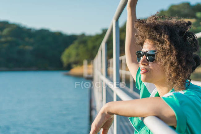 Ritratto di donna con occhiali da sole appoggiata alla ringhiera del molo sul lago illuminato dal sole — Foto stock