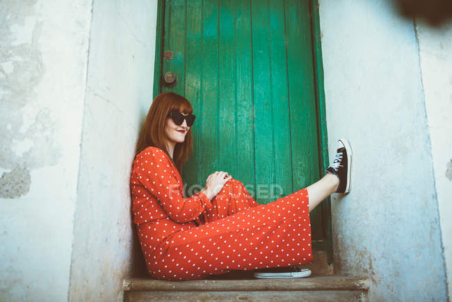 Рыжая женщина в красном платье и солнечных очках сидит на пороге — стоковое фото