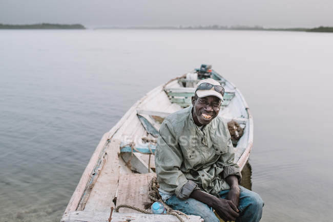 Yoff, Senegal- 6 dicembre 2017: Ritratto di uomo sorridente che guarda la macchina fotografica e siede su una vecchia barca a vela sulla riva del fiume . — Foto stock