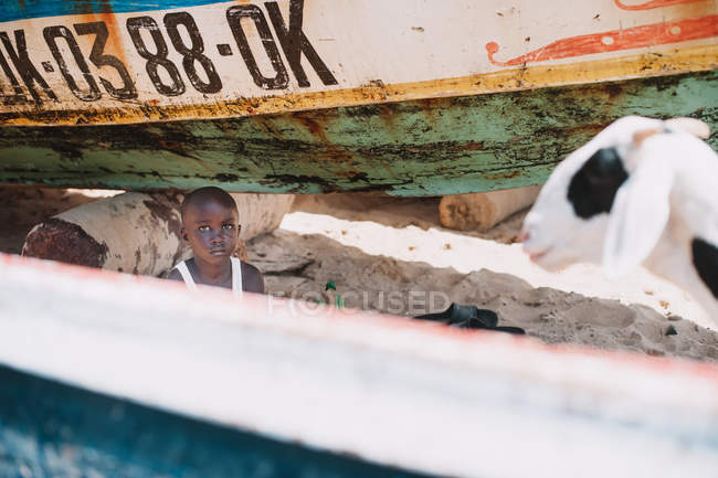 Yoff, Senegal- 6 de diciembre de 2017: Retrato de un niño mirando a la cámara y sentado en vasos con cabra  . - foto de stock