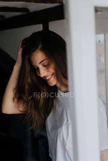 Портрет улыбающейся брюнетки в повседневной футболке, касающейся волос и смотрящей вниз у окна — стоковое фото