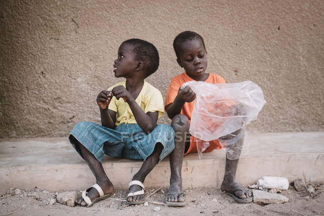 Yoff, Senegal- December 6, 2017: Two little boys sitting with plastic bag on grungy sidewalk. — Stock Photo