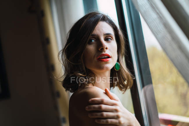 Chica morena con labios rojos abrazando hombro y posando cerca de la ventana - foto de stock