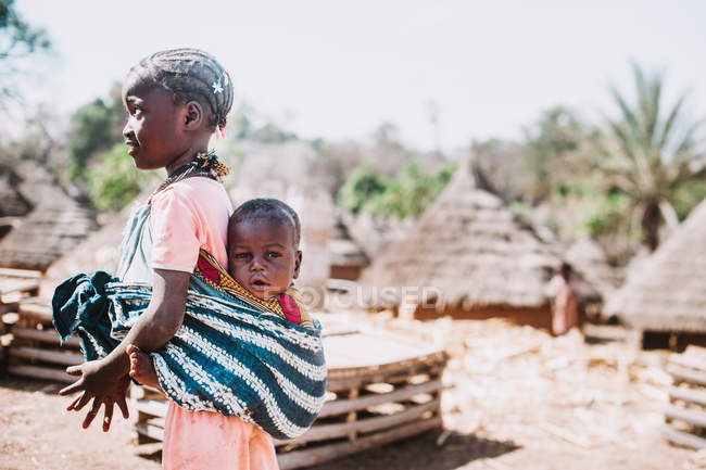 Кедугу, Сенегал - 8 декабря 2017 г.: Вид сбоку на девочку с ребенком на спине в племенной деревне — стоковое фото