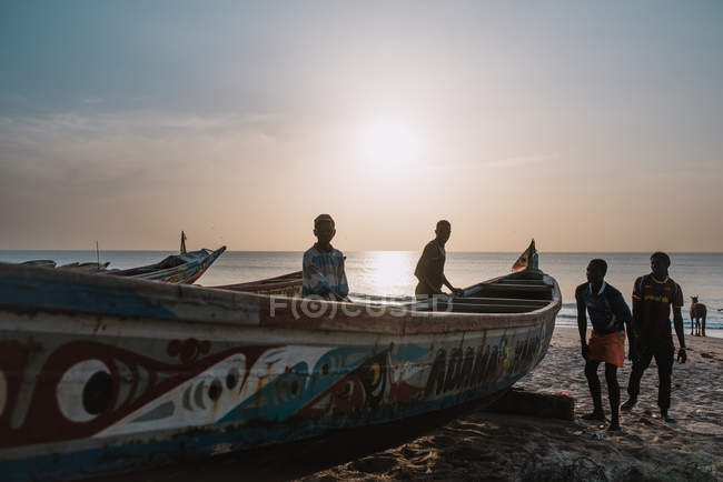 Goree, Senegal- 6 de diciembre de 2017: Grupo de adolescentes africanos de pie en la playa cerca del barco al atardecer . - foto de stock