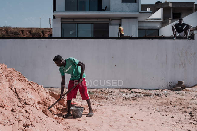 Goree, Senegal- December 6, 2017: Black worker in bright sunlight loading sand in bucket with shovel. — Stock Photo
