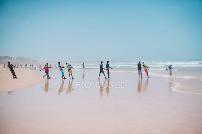 Goree, Senegal - 6 de diciembre de 2017: Fila de personas de pie en la orilla del mar y tirando de la cuerda en la playa de arena . - foto de stock