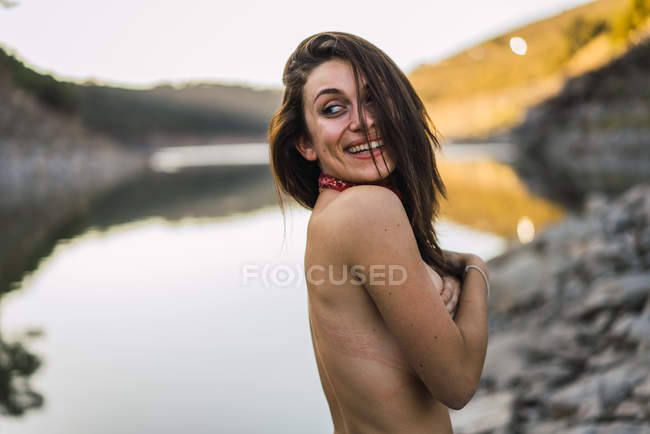 Vue latérale d'une jeune brune cachant sa poitrine sur fond de lac de montagne l — Photo de stock