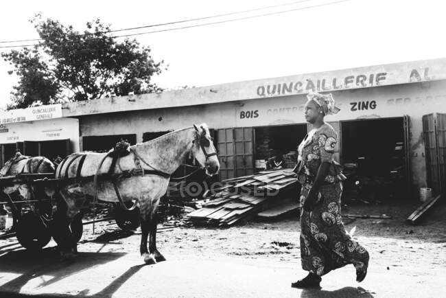 Африканская женщина в традиционной одежде идет по улице рядом с лошадью. — стоковое фото